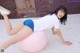 A woman in a white shirt and blue shorts is sitting on a pink ball.